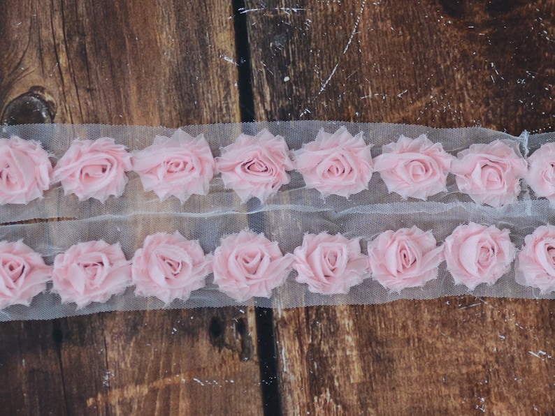 May include: Pink fabric rose trim on a white mesh background. The trim is made of multiple layers of fabric to create a 3D effect.
