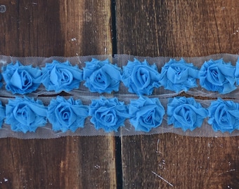 May include: Blue fabric flower trim on a white mesh background. The trim is made of small, ruffled fabric flowers.