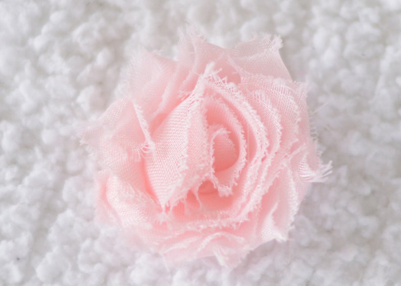 May include: A delicate pink fabric flower with ruffled edges. The flower is made of soft, sheer fabric and has a layered, rosette-like design.