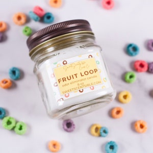 May include: A clear glass jar candle with a brown lid. The label reads "Sweet Scents-ible" and "Fruit Loop odor eliminator candle 8 oz SWEETSCENTSIBLE.COM". The candle is surrounded by colorful cereal rings.