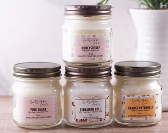 May include: Four glass jars with lids containing candles. The candles are labeled with the scents: Honeysuckle, Pink Sugar, Cinnamon Roll, and Orange Patchouli. Each jar is 8 oz.