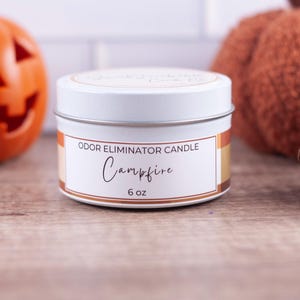 May include: A white tin candle with a black and orange label that reads "ODOR ELIMINATOR CANDLE Campfire 6 oz".