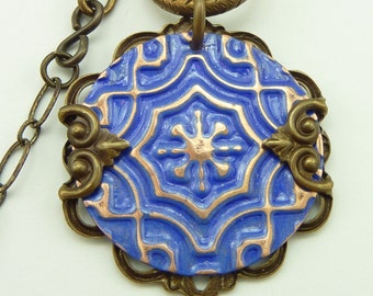 May include: A blue and copper pendant necklace with a detailed geometric design. The pendant is attached to a brass chain with a clasp.