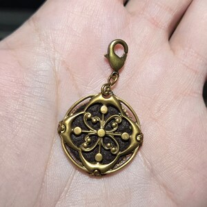 May include: A gold-toned charm with a detailed floral design. The charm is attached to a metal loop and has a dark brown center.