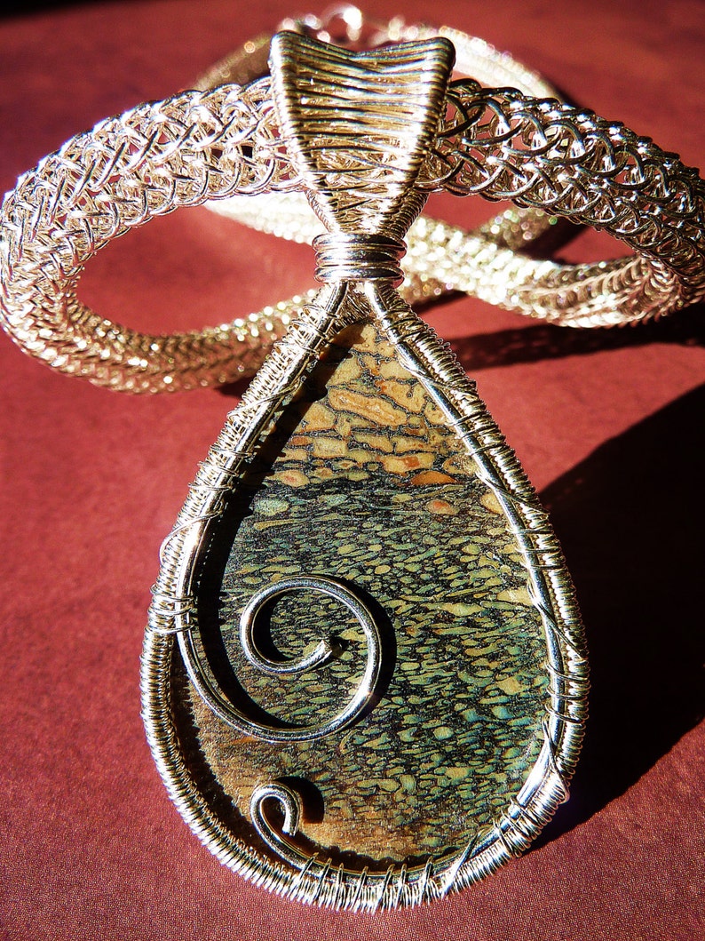 May include: A silver wire wrapped pendant featuring a fossilized stone with a swirling pattern. The stone is a deep green and brown with a light brown border. The pendant is suspended from a silver chain.