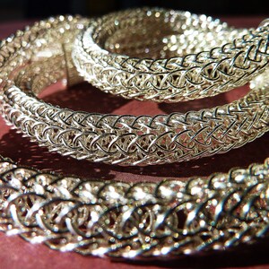 May include: A silver chainmail necklace with a woven design. The necklace is made of small, interconnected metal rings.