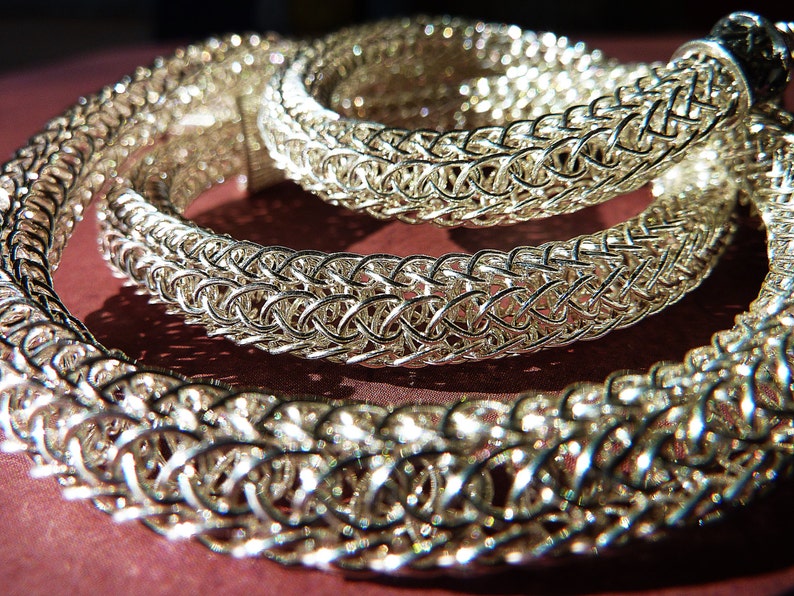 May include: A silver chainmail necklace with a woven design. The necklace is made of small, interconnected metal rings.