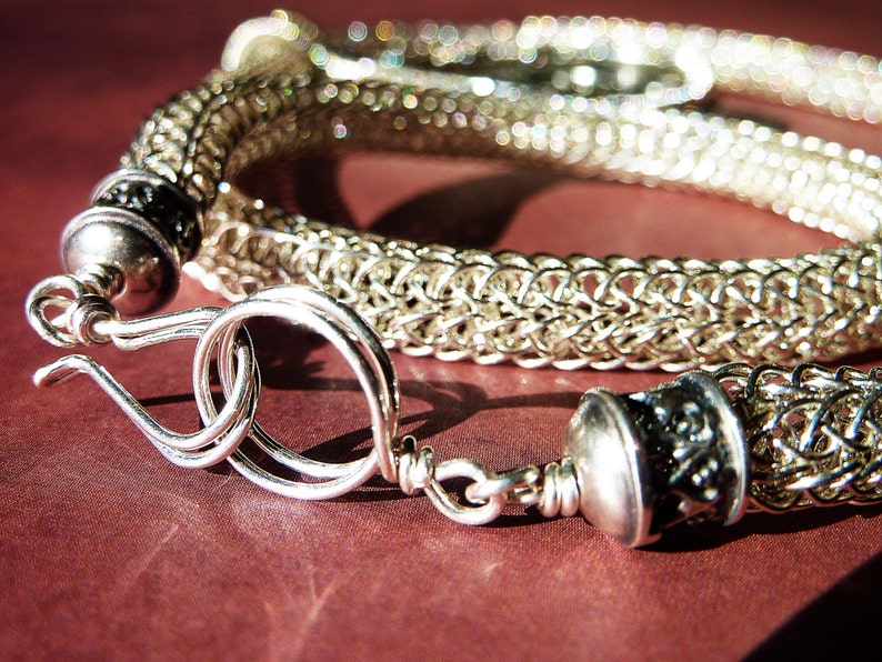 May include: A silver chainmail bracelet with a decorative clasp. The bracelet is made of small, interconnected metal rings.