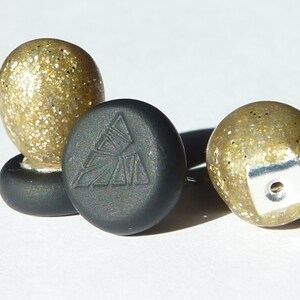 Può includere: Un paio di orecchini con glitter dorati e retro nero. Gli orecchini hanno un design geometrico sul retro.