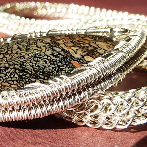 May include: A silver wire wrapped pendant featuring a black and white patterned stone. The pendant is attached to a silver chain.