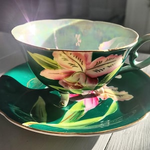 May include: A vintage teacup and saucer set. The cup and saucer are a deep green with a floral design in pink and yellow. The cup has a unique handle and sits on three small feet. The rim of the cup has a pearlescent sheen.