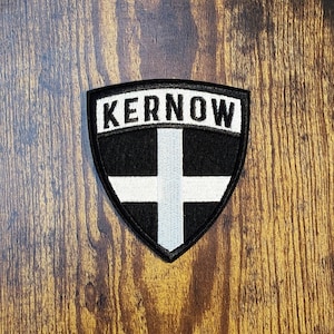 May include: A shield-shaped embroidered patch featuring a black background with a white cross design. The word KERNOW is stitched in bold white capital letters across the top section of the patch, which is bordered by a thick black stitched edge.