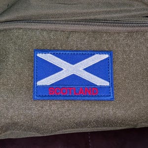May include: A rectangular embroidered patch featuring a blue background with a white diagonal cross and the word SCOTLAND stitched in red capital letters at the bottom. The patch is attached to a dark olive green fabric surface with a zipper.