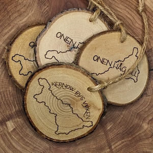 May include: Four wooden ornaments with a natural bark edge and a jute string for hanging. Each ornament features a map outline and text, including "ONEN HÅG" and "KERNOW BYS VYKEN" etched into the wood. The ornaments are arranged on a wooden surface.