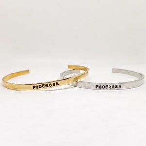 May include: Two open cuff bracelets. One is gold-toned, the other is silver-toned. Both have the word "PODEROSA" engraved in black, centered on the front. The bracelets are displayed on a white background.