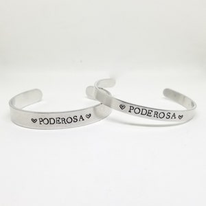 May include: Two silver bracelets with the word "PODEROSA" engraved on them, each with a heart symbol on either side of the word.
