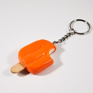 May include: Orange popsicle keychain with a bite taken out of it. The popsicle is attached to a silver key ring.