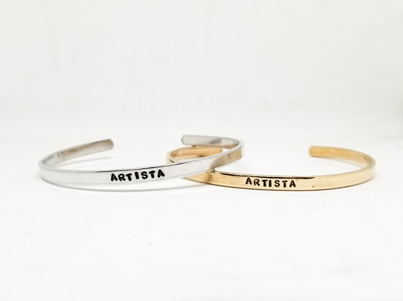 May include: Two open cuff bracelets, one silver-toned and one gold-toned, are displayed against a white background. The word "ARTISTA" is stamped in black on the front of each bracelet. These bracelets are a simple and elegant accessory.