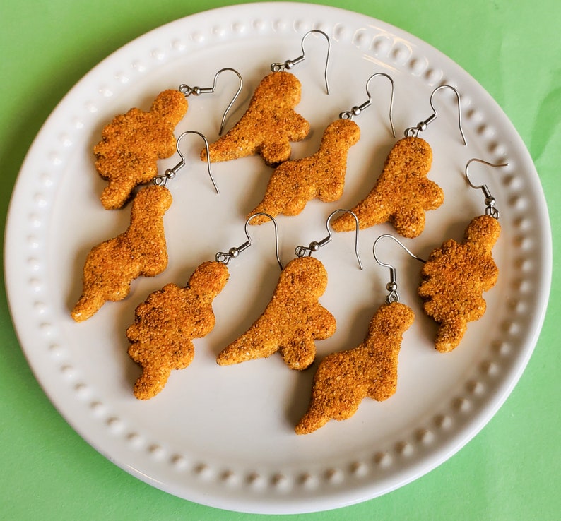 May include: A set of eight dinosaur-shaped earrings made of brown and tan cookie dough. The earrings are displayed on a white plate with a scalloped edge.
