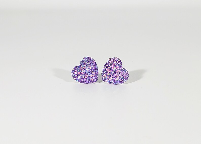 K&ouml;nnte beinhalten: Ein Paar violette herzf&ouml;rmige Ohrringe mit schimmerndem Glitzer.