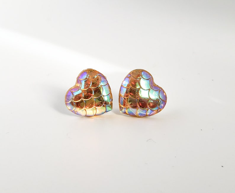 Puede incluir: Un par de pendientes de bot&oacute;n en forma de coraz&oacute;n con un dise&ntilde;o iridiscente en forma de escamas. Los pendientes presentan una mezcla de tonos dorados, anaranjados y azules, creando un efecto brillante. Los pendientes est&aacute;n sobre un fondo blanco.