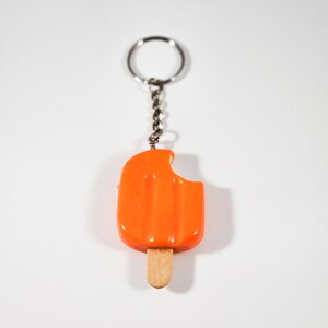 May include: Orange popsicle keychain with a bite taken out of it. The popsicle is attached to a silver chain and ring.