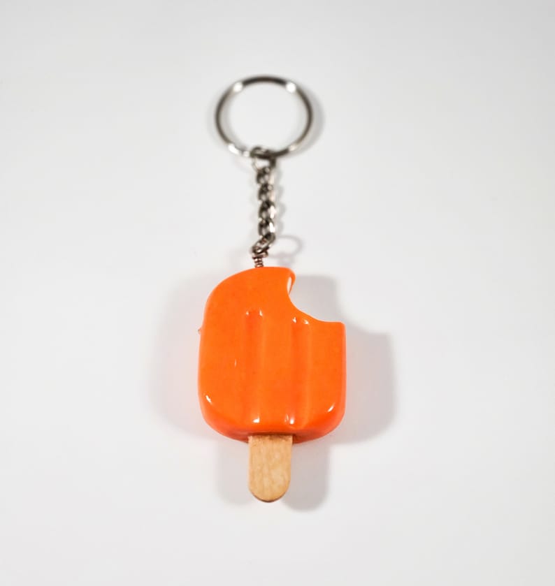May include: Orange popsicle keychain with a bite taken out of it. The popsicle is attached to a silver chain and ring.