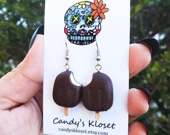 May include: A pair of dangle earrings featuring a chocolate-covered ice cream bar with a bite taken out. The earrings are on a white card with a colorful sugar skull illustration and the text "Candy's Kloset" and "candyskloset.etsy.com".