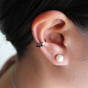 Peut inclure: Une personne portant une petite boucle d'oreille en perle et une manchette d'oreille en argent avec trois pierres noires.