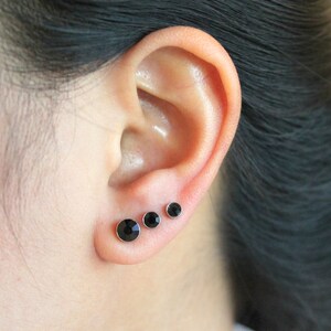 May include: Close-up of a person's ear with three black stud earrings. The earrings are of different sizes and are arranged in a row.