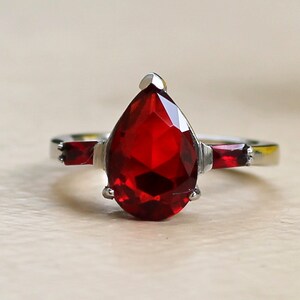 Peut inclure: Une bague en argent avec une grande pierre rouge en forme de poire et deux petites pierres rouges sur la bande.