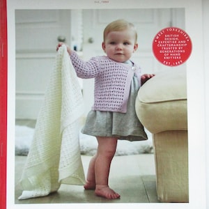 Peut inclure: Un bébé portant un cardigan violet clair et une robe grise, tenant une couverture blanche tricotée. L'image comprend le texte "Sirdar" et "Snuggly 100% Merino". Le modèle est pour les 0-3 ans.