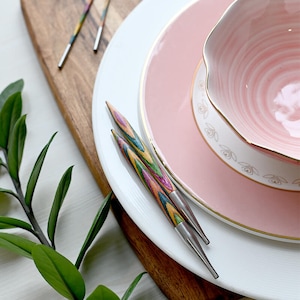 Peut inclure: Deux aiguilles à tricoter en bois colorées avec des pointes métalliques sont sur une assiette blanche avec une assiette rose et un bol rose sur le dessus. Les assiettes et le bol ont une bordure dorée.