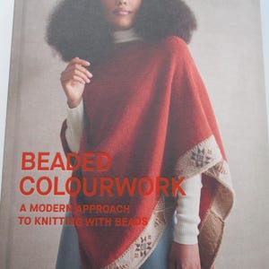 May include: A book titled Beaded Colourwork: A Modern Approach to Knitting with Beads by Jeanette Sloan. The cover features a person wearing a rust-coloured knitted poncho with a cream-coloured beaded border over a white ribbed sweater and blue skirt.