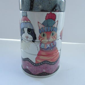 May include: A multi-tiered ceramic container with a lid, featuring illustrations of cats in winter attire. The cats, in shades of black, white, and orange, are depicted wearing hats and scarves. The lid has a speckled grey and blue design.