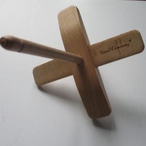 以下が含まれることがあります： 木製の糸巻き機。糸をかせに巻くための道具です。明るい色の木材で作られており、中央にダボが付いた十字型のベースがあります。「Hand U Journey」の文字が腕の1つに見えます。