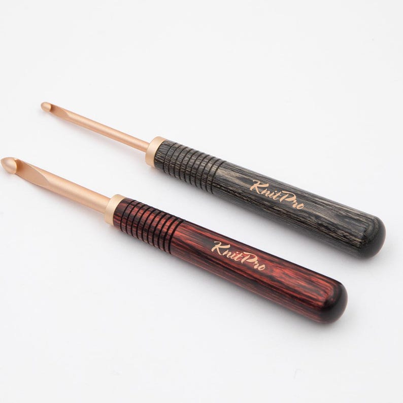May include: Two wooden crochet hooks with rose gold tips. The hooks are made of wood with a dark brown and a reddish brown finish. The hooks are labeled "KnitPro".