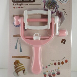 May include: A pink and white pom pom and tassel rolling maker on a white background. The package includes text that says "Pom Pom and Tassel Rolling Maker" and "Just turn the handle around and enjoy!"