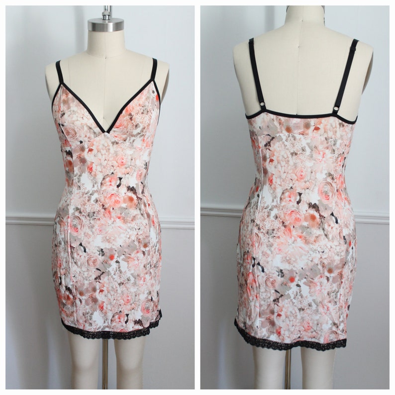 May include: A white slip dress with a floral print in shades of pink and orange. The dress has a V-neckline and thin black straps. The hemline is trimmed with black lace.