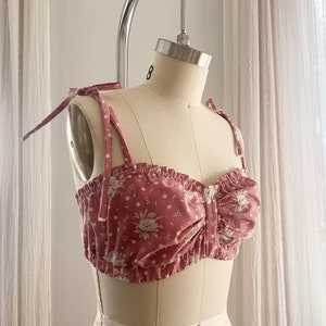 May include: A pink floral print crop top with adjustable straps. The top has a gathered detail in the front and a ruched design.