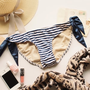 Puede incluir: Braga de bikini a rayas azules y blancas con un lazo azul marino a cada lado. La braga de bikini está sobre una superficie blanca con un sombrero de paja, una bufanda de estampado de leopardo y otros artículos.