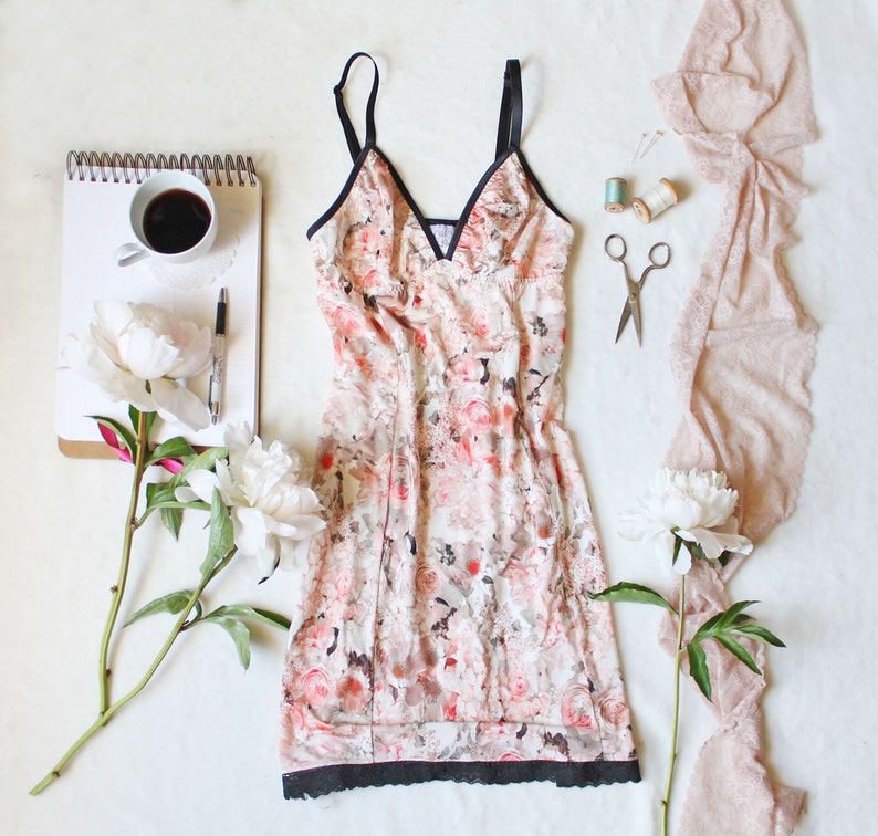 May include: A floral print slip dress with a black lace trim. The dress is made of a soft, lightweight fabric and has a relaxed fit. The dress is photographed on a white background with flowers and a notebook.