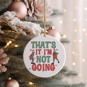 May include: White ceramic Christmas ornament with a green and red design. It features two cartoon figures wearing Santa hats and the text "That's it I'm not going."