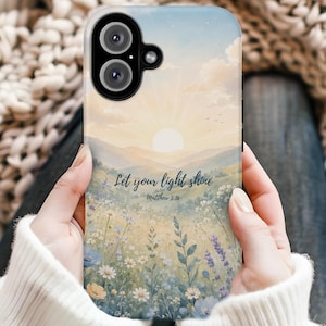 May include: A phone case featuring a watercolor-style landscape of a sunrise over rolling hills and a field of wildflowers. The text 'Let your light shine' and 'Matthew 5:16' is printed in dark blue script in the center of the design.