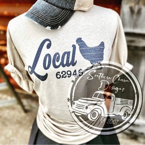 May include: A beige t-shirt with a blue graphic of a chicken and the text "Local 62946". The shirt has a distressed look and is paired with a denim cap. The image also includes a logo for Southern Charm Designs, featuring a vintage truck and the text "Southern Charm Designs Est 2007".