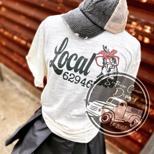 May include: A white t-shirt with the text "Local 62946" and a cartoon chicken wearing glasses and a red bandana. The shirt is worn by a person wearing a black skirt and a gray and beige baseball cap. The image also includes a logo for "Southern Charm Designs Est. 2007" with a vintage truck.