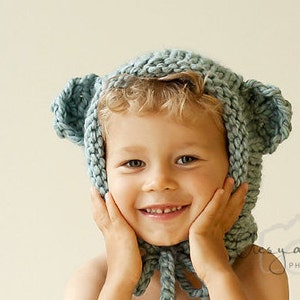 May include: A child wearing a light blue knitted hat with bear ears. The hat has a long, flowing scarf attached to it. The child is smiling and has their hands on their cheeks.