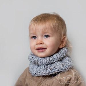 May include: A young child with blonde hair wearing a grey knitted cowl neck scarf. The child is looking at the camera and smiling.
