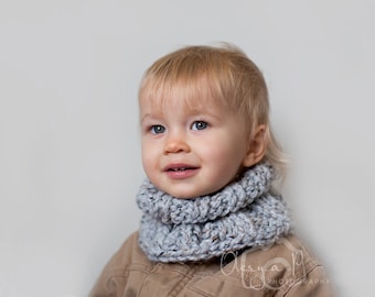 May include: A young child with blonde hair wearing a grey knitted cowl neck scarf. The child is looking at the camera and smiling.