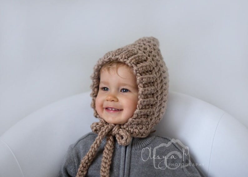 K&ouml;nnte beinhalten: Ein Baby tr&auml;gt eine braune gestrickte Kapuze mit einer langen Schleife. Die M&uuml;tze hat ein strukturiertes Stichmuster.